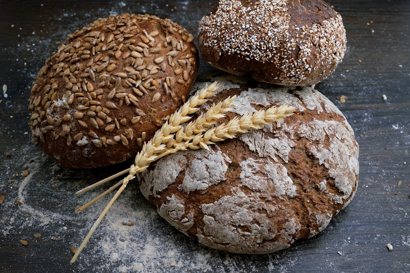 Three rustic loaves of bread and wheat stalks on a wooden table.