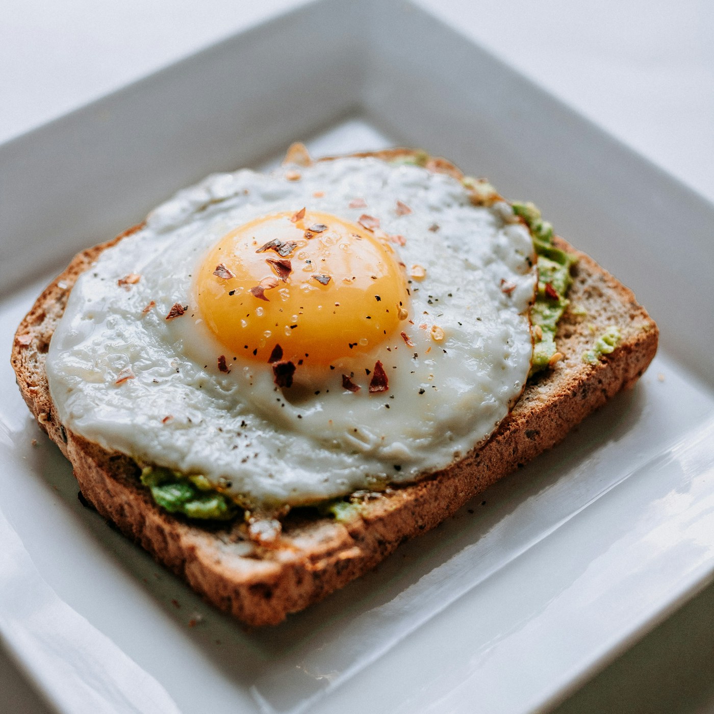 A fried egg on avocado toast, seasoned with pepper and chili.