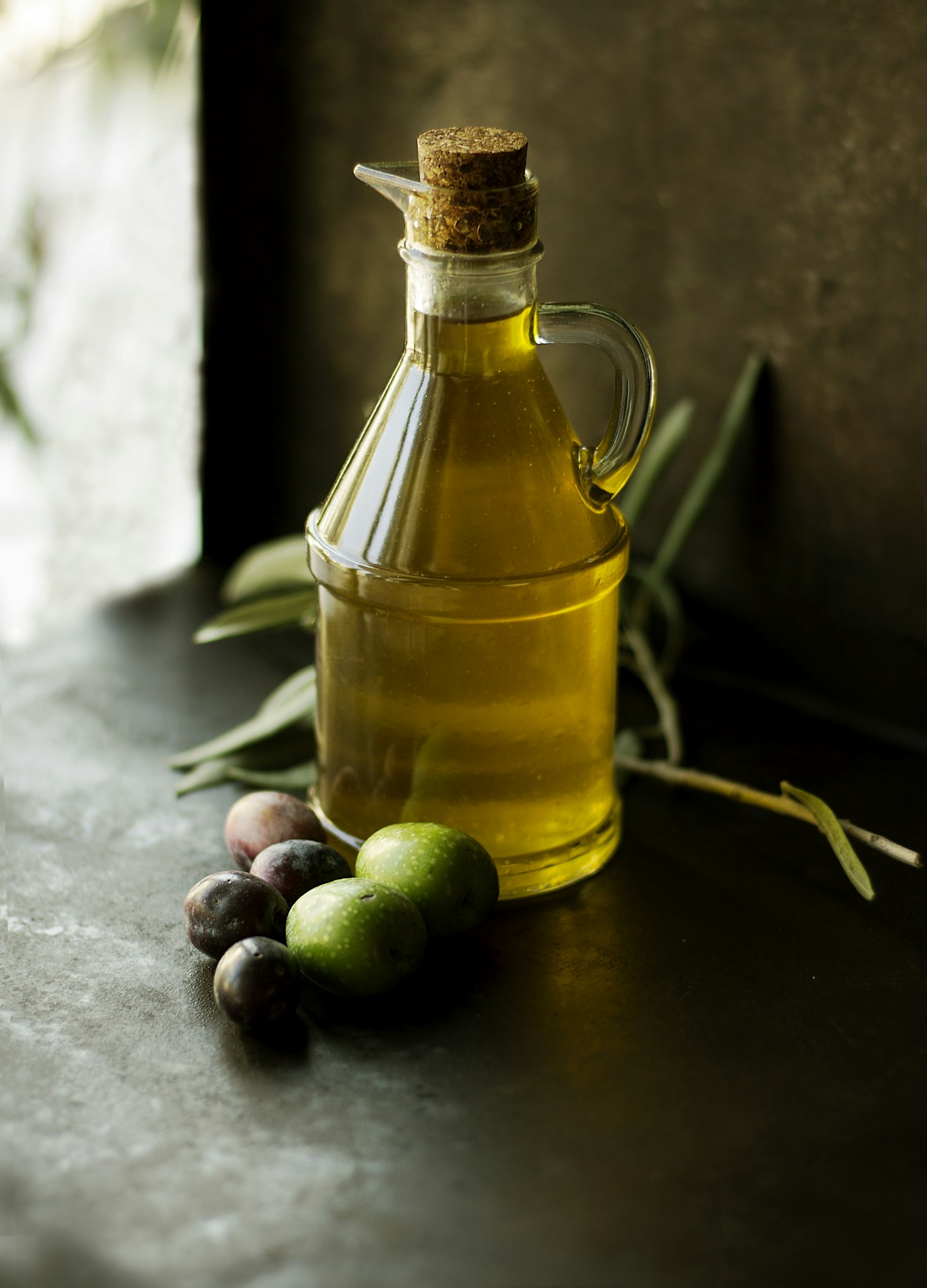 A glass carafe of olive oil with olives and a branch on a dark surface.